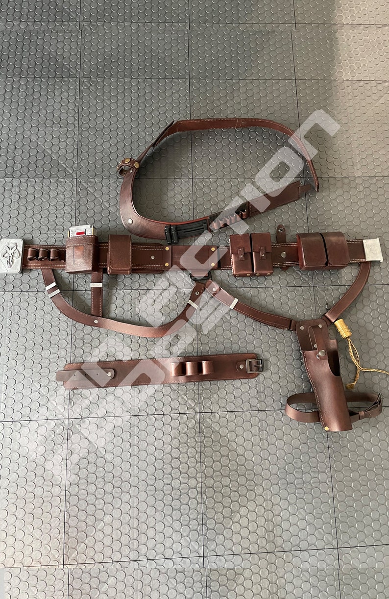May include: A brown leather belt with multiple pouches and straps. The belt has a silver buckle and a silver skull emblem. The belt is laid out on a gray textured surface.