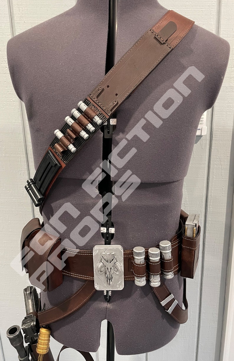 May include: A brown leather belt with a silver buckle featuring a Mandalorian symbol. The belt has a brown leather holster on the left side and a pouch on the right side. There are also several small silver tubes attached to the belt. A brown leather strap with a black buckle is worn diagonally across the chest.