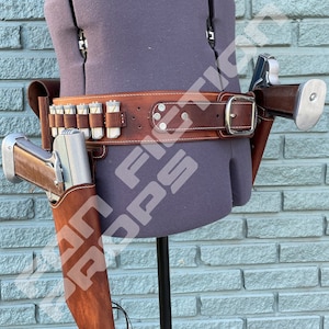 May include: A brown leather belt with a silver buckle and two holsters for pistols. The belt has a row of bullet loops and is worn on a mannequin.