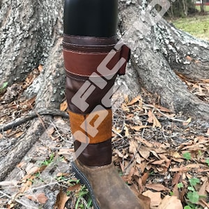 May include: Brown leather riding boots with a black top and brown leather straps. The boots are sitting on a bed of brown leaves and green grass.