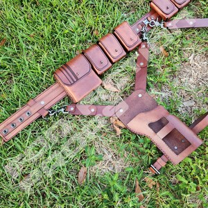 Puede incluir: Un cintur&oacute;n utilitario de cuero marr&oacute;n con m&uacute;ltiples bolsillos y una funda, sobre hierba verde. El cintur&oacute;n presenta herrajes met&aacute;licos y est&aacute; dise&ntilde;ado para transportar varios objetos. El cuero tiene un aspecto rico y texturizado.