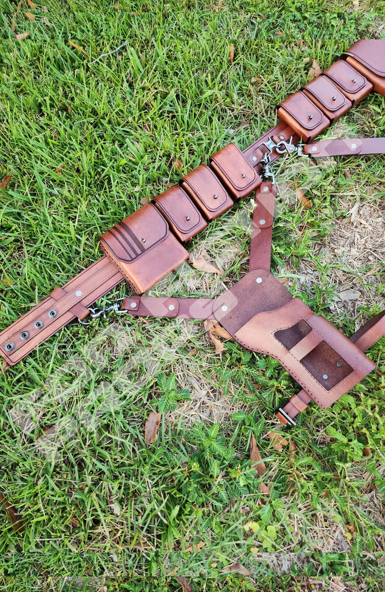 Puede incluir: Un cintur&oacute;n utilitario de cuero marr&oacute;n con m&uacute;ltiples bolsillos y una funda, sobre hierba verde. El cintur&oacute;n presenta herrajes met&aacute;licos y est&aacute; dise&ntilde;ado para transportar varios objetos. El cuero tiene un aspecto rico y texturizado.