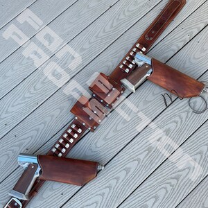 May include: A brown leather gun belt with two holsters and a cartridge belt. The belt has silver studs and is laid out on a wooden surface.
