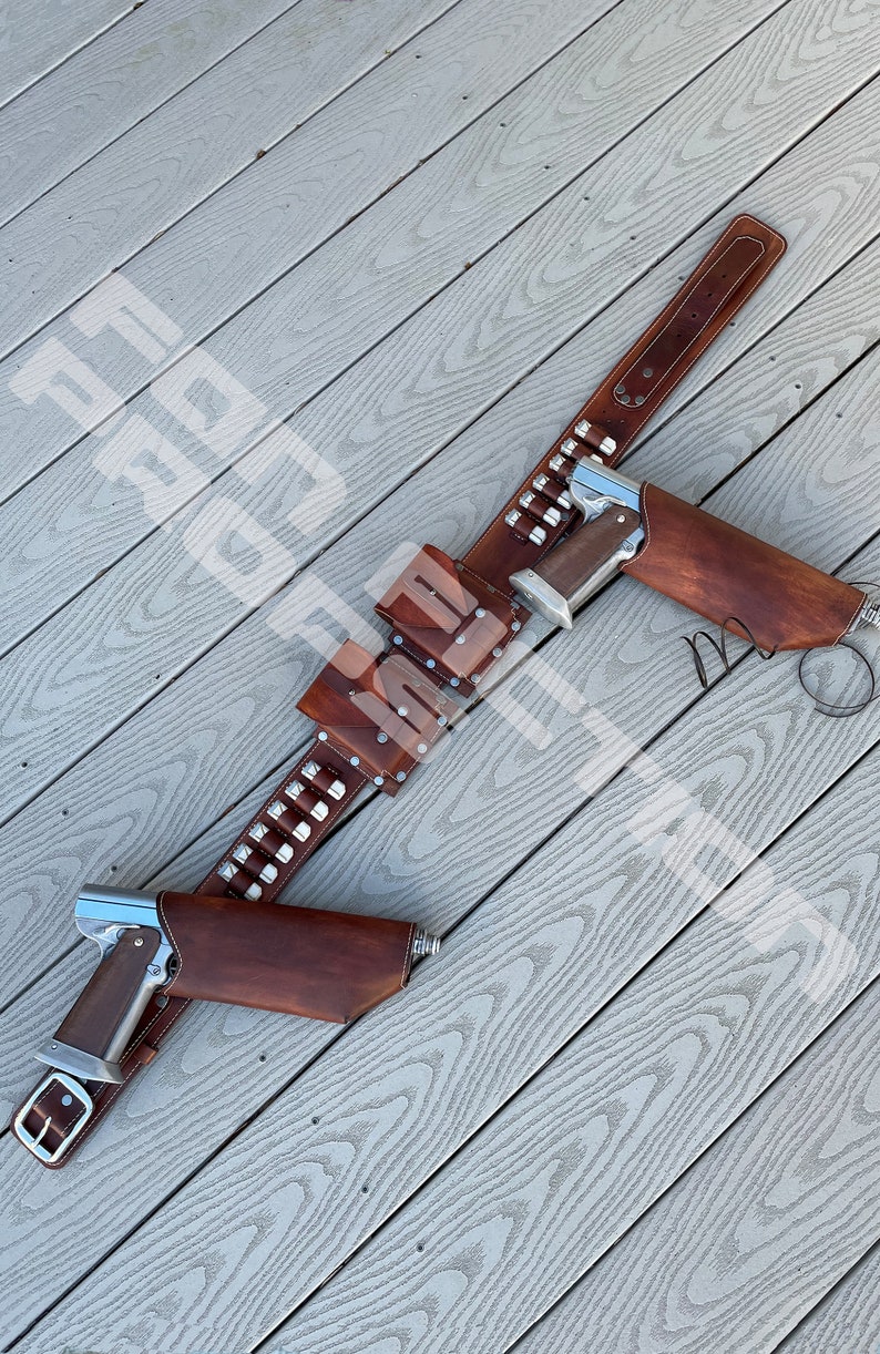 May include: A brown leather gun belt with two holsters and a cartridge belt. The belt has silver studs and is laid out on a wooden surface.