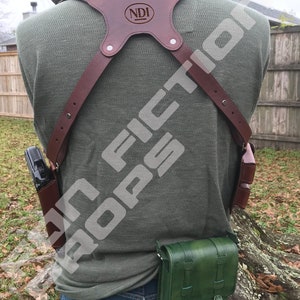 May include: A brown leather holster with a green leather pouch attached to it. The holster has a strap that goes over the shoulder and a buckle on the chest. The pouch has two pockets with buckles. The holster is labelled "NDI".