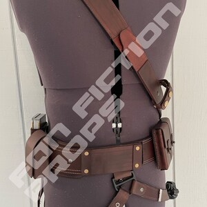 May include: A brown leather belt with a holster and a shoulder strap. The belt has a buckle and multiple brass rivets. The holster has a flap and a strap. The shoulder strap has a buckle and a loop.