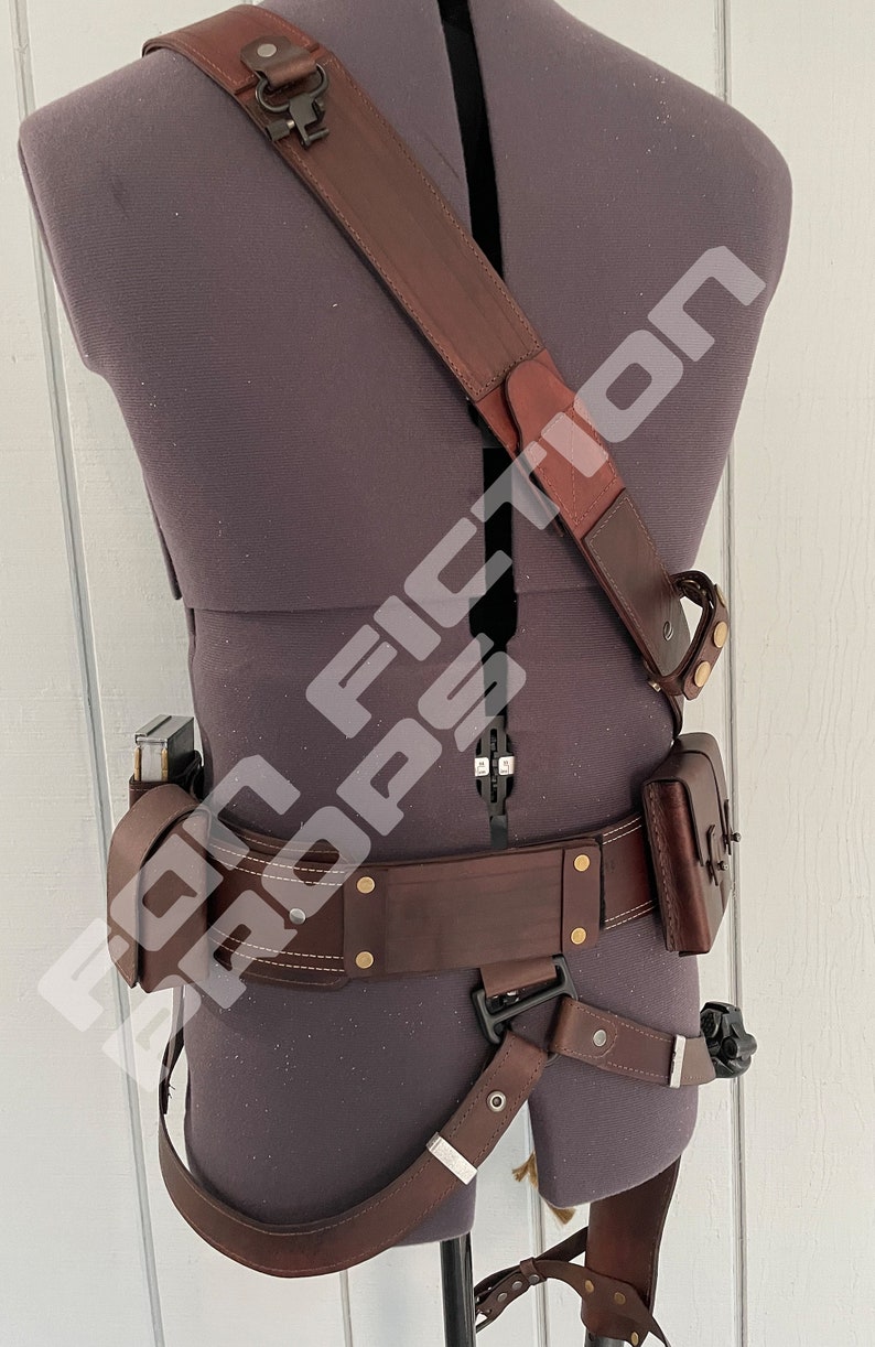 May include: A brown leather belt with a holster and a shoulder strap. The belt has a buckle and multiple brass rivets. The holster has a flap and a strap. The shoulder strap has a buckle and a loop.