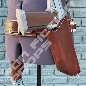 May include: A silver and brown pistol with a brown leather holster. The holster is attached to a brown leather belt with a silver buckle. The pistol has a black cord attached to it.