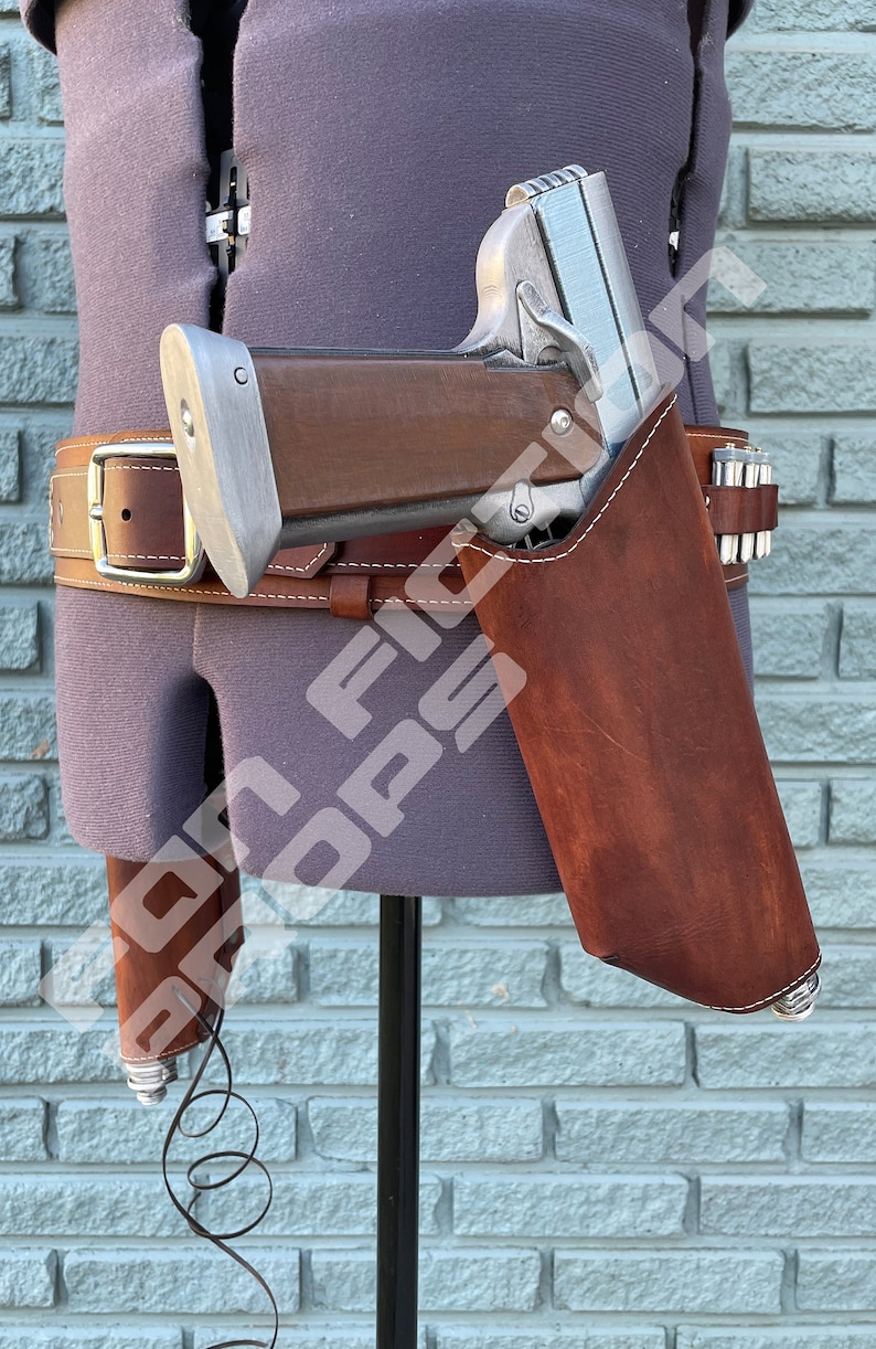 May include: A silver and brown pistol with a brown leather holster. The holster is attached to a brown leather belt with a silver buckle. The pistol has a black cord attached to it.