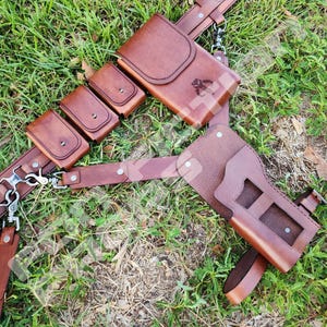 Puede incluir: Un cintur&oacute;n utilitario de cuero marr&oacute;n con m&uacute;ltiples bolsillos y una funda. El cintur&oacute;n tiene un cierre de hebilla y remaches met&aacute;licos. El dise&ntilde;o incluye un s&iacute;mbolo en el bolsillo m&aacute;s grande. El cintur&oacute;n est&aacute; sobre hierba verde.