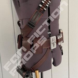 May include: A brown leather belt with a silver buckle featuring a Mandalorian symbol. The belt has a holster for a gun and a pouch for carrying supplies. The belt is worn on a mannequin.