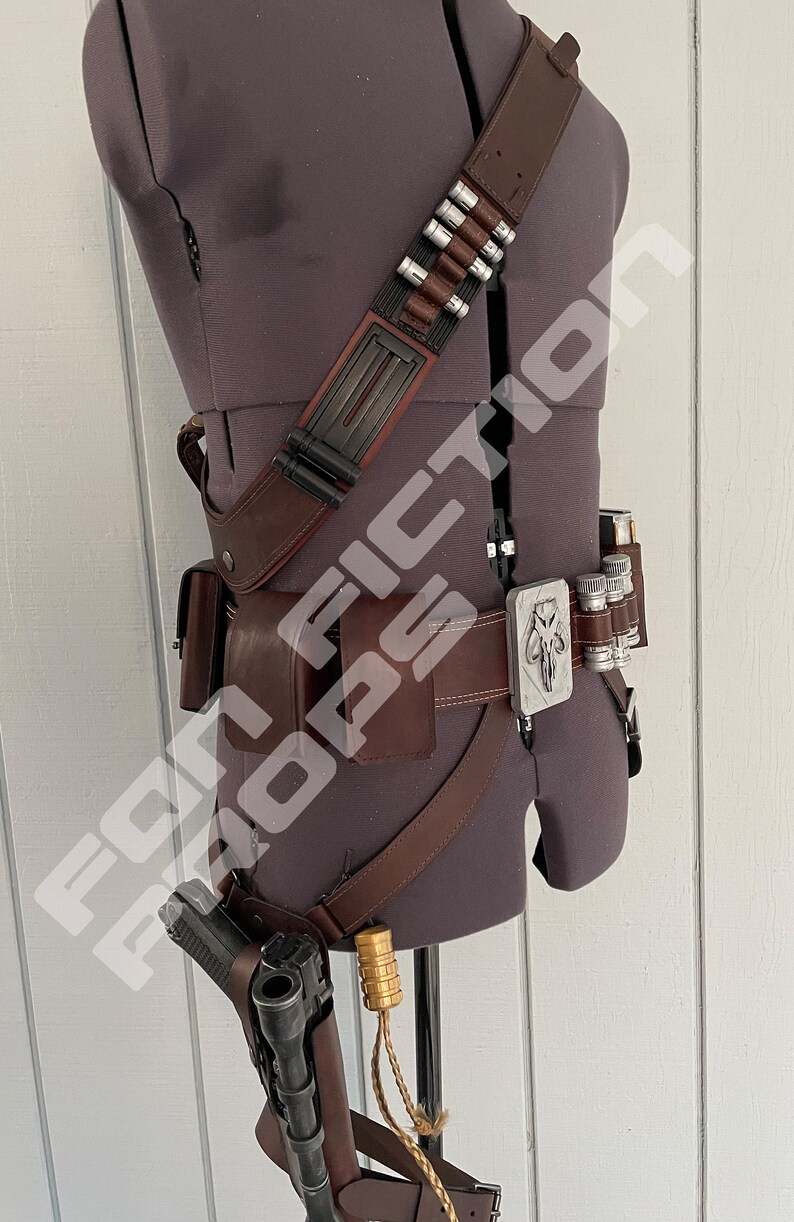 May include: A brown leather belt with a silver buckle featuring a Mandalorian symbol. The belt has a holster for a gun and a pouch for carrying supplies. The belt is worn on a mannequin.