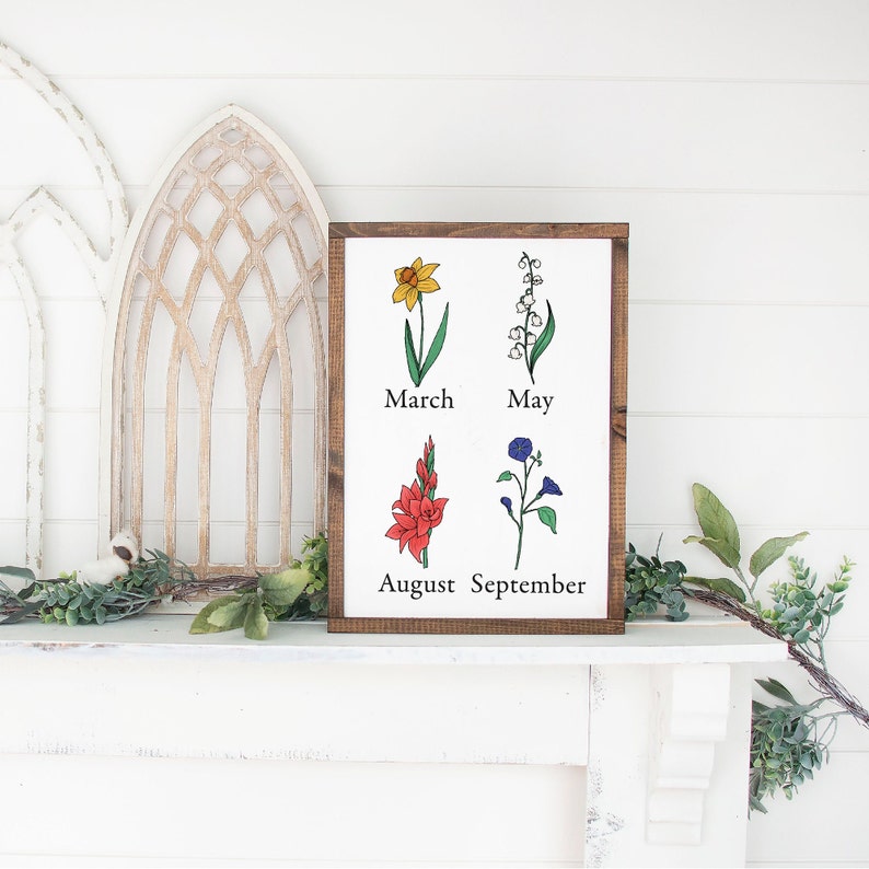 May include: A wooden sign with a white background and brown frame. The sign features illustrations of flowers and the months they bloom: March (daffodil), May (lily of the valley), August (gladiolus), and September (morning glory).