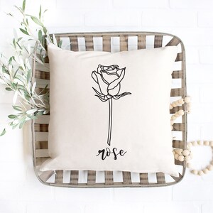 May include: A white throw pillow with a black line drawing of a rose and the word "rose" written in cursive below the rose.