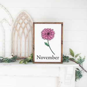 May include: A wooden sign with a brown frame and a white background. The sign features a pink flower with green leaves and the word "November" in black text.