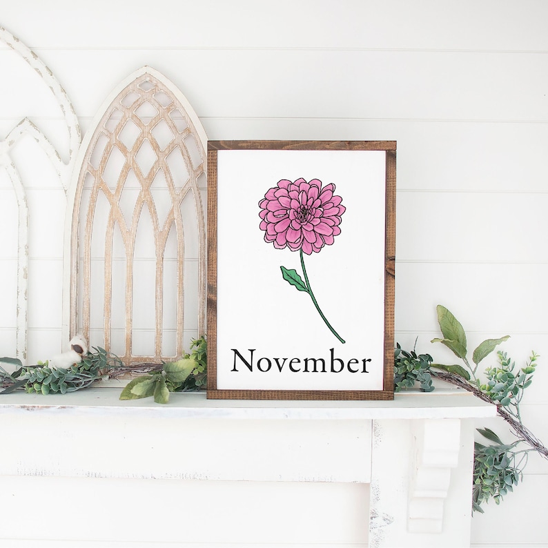 May include: A wooden sign with a brown frame and a white background. The sign features a pink flower with green leaves and the word "November" in black text.