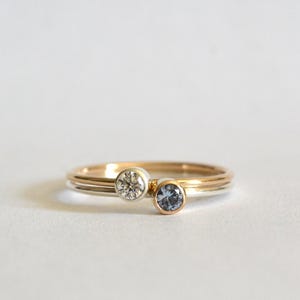 May include: Two delicate gold rings, one with a clear gemstone and the other with a blue gemstone. The rings are stacked, showcasing a minimalist design. The gemstones are round and set in a bezel setting.