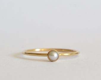 May include: A delicate gold ring featuring a single, round, white pearl. The ring has a thin band and a simple, elegant design. The pearl is set in a gold bezel.