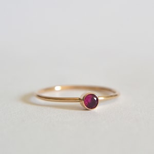 May include: A simple gold band ring with a single, small, round, red ruby gemstone.