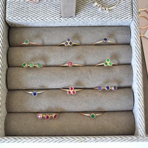 K&ouml;nnte beinhalten: Nahaufnahme eines Schmuckauslagenkastens mit einer Auswahl an goldenen Ringen mit verschiedenen farbigen Edelsteinen. Die Ringe sind in Reihen auf einem beigefarbenen Samtstoff angeordnet. Die Edelsteine umfassen Gr&uuml;n, Blau und Rot.