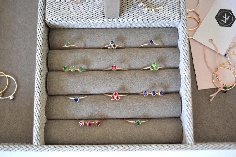 K&ouml;nnte beinhalten: Nahaufnahme eines Schmuckauslagenkastens mit einer Auswahl an goldenen Ringen mit verschiedenen farbigen Edelsteinen. Die Ringe sind in Reihen auf einem beigefarbenen Samtstoff angeordnet. Die Edelsteine umfassen Gr&uuml;n, Blau und Rot.