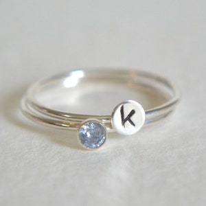 May include: Two silver rings, one with a small blue gemstone and the other with the letter "K" engraved on it.