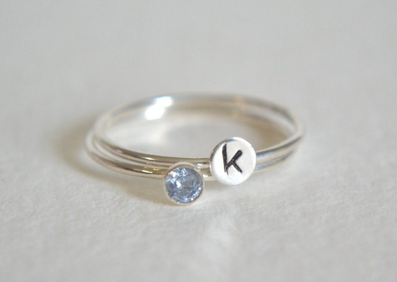 May include: Two silver rings, one with a small blue gemstone and the other with the letter "K" engraved on it.