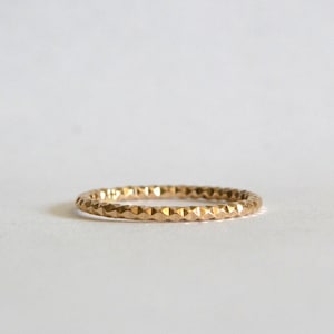 Può includere: Un anello color oro con un motivo geometrico strutturato. L'anello è sottile e sembra essere in metallo. Il design presenta una serie di piccole forme ripetute attorno alla fascia. L'anello è posto su uno sfondo bianco.