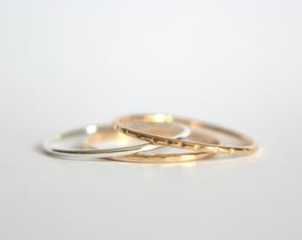 May include: Three thin gold and silver rings stacked on top of each other. The top ring is gold with a textured surface. The middle ring is silver and the bottom ring is gold with a smooth surface.