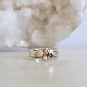May include: A set of four silver stacking rings with a white opal, a clear diamond, a pearl, and a blue topaz gemstone.