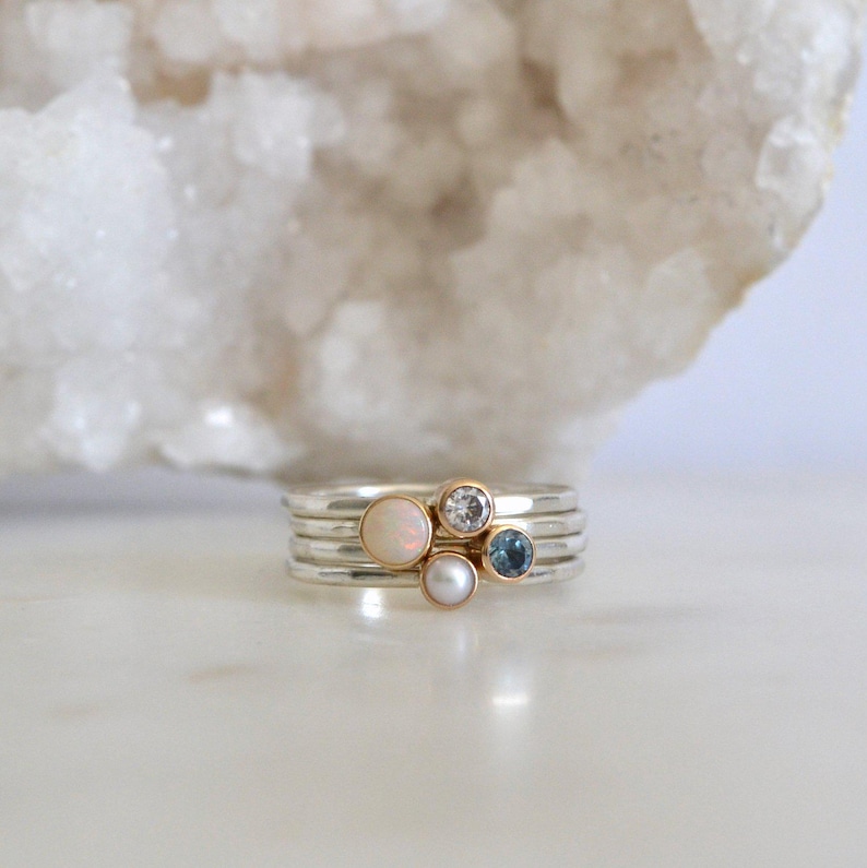 May include: A set of four silver stacking rings with a white opal, a clear diamond, a pearl, and a blue topaz gemstone.