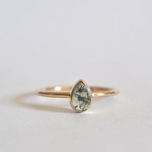 May include: A gold ring with a teardrop-shaped moss agate gemstone. The gemstone is a light green and white color with a swirling pattern.