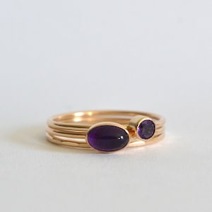May include: A set of three gold rings with amethyst gemstones. The rings are stacked on top of each other, with the largest ring featuring a larger oval-shaped amethyst and the smaller rings featuring round amethysts.