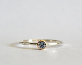 May include: A delicate ring with a silver band and a gold bezel setting. The ring features a small, round, blue gemstone. The band has a hammered texture, adding a subtle detail to the minimalist design.