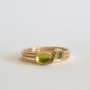 Puede incluir: Un conjunto de tres anillos de oro con piedras preciosas verdes. Los anillos están apilados uno encima del otro, con el anillo más grande en la parte inferior y el anillo más pequeño en la parte superior.