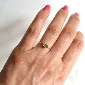 Puede incluir: Un delicado anillo de oro con una peque&ntilde;a piedra preciosa verde de forma ovalada. El anillo se lleva en un dedo.