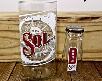 May include: Two clear glass bottles with the Sol Cerveza logo. The larger bottle has the text "Desde 1899" and "Original de Mexico". The smaller bottle has the text "Desde 1899" and "Sol".