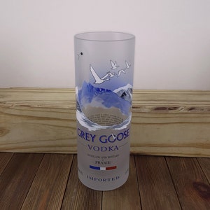 May include: A clear glass Grey Goose Vodka bottle repurposed as a drinking glass. The glass features a frosted design with a blue mountain scene and flying geese. The text "GREY GOOSE VODKA" is printed in blue on the glass. The text "DISTILLED AND BOTTLED IN FRANCE" is printed in blue on the glass. The French flag is printed in blue, white, and red on the glass. The text "IMPORTED" is printed in blue on the glass.