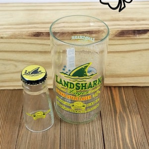 May include: A clear glass with a Land Shark Island Style Lager label. The label features a green shark fin and the text "Land Shark Lager" in yellow and green. The glass is sitting on a wooden surface next to a clear glass bottle with a yellow and black cap.