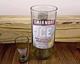 Puede incluir: Una botella de vidrio transparente con una etiqueta de Smirnoff Ice Original. La botella está sobre una superficie de madera junto a un vaso de chupito transparente con una etiqueta de Smirnoff Ice Original.