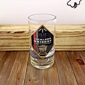 May include: A clear glass with a label that reads "52 Distilling Cackler's Whiskey A Blend of Rye Bourbon Whiskey" with a gold and black design. The label also includes the text "Est. 2017" and "750 ml - 40% Alc/Vol".