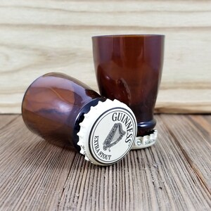 May include: Two brown glass shot glasses with a Guinness bottle cap on the bottom of each. The bottle caps are white with black lettering that reads "Guinness Extra Stout".