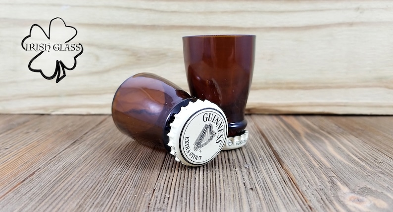 May include: Two brown glass shot glasses with a Guinness bottle cap on the bottom of each. The bottle caps are white with black lettering that reads "Guinness Extra Stout".