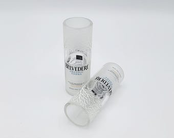 Könnte beinhalten: Zwei klare Trinkgläser, die aus Belvedere Organic Vodka Flaschen gefertigt wurden. Jedes Glas zeigt ein weißes, geätztes Baumzweig-Design und das in Schwarz gedruckte Belvedere Organic Vodka Logo. Der obere Rand ist glatt und klar.