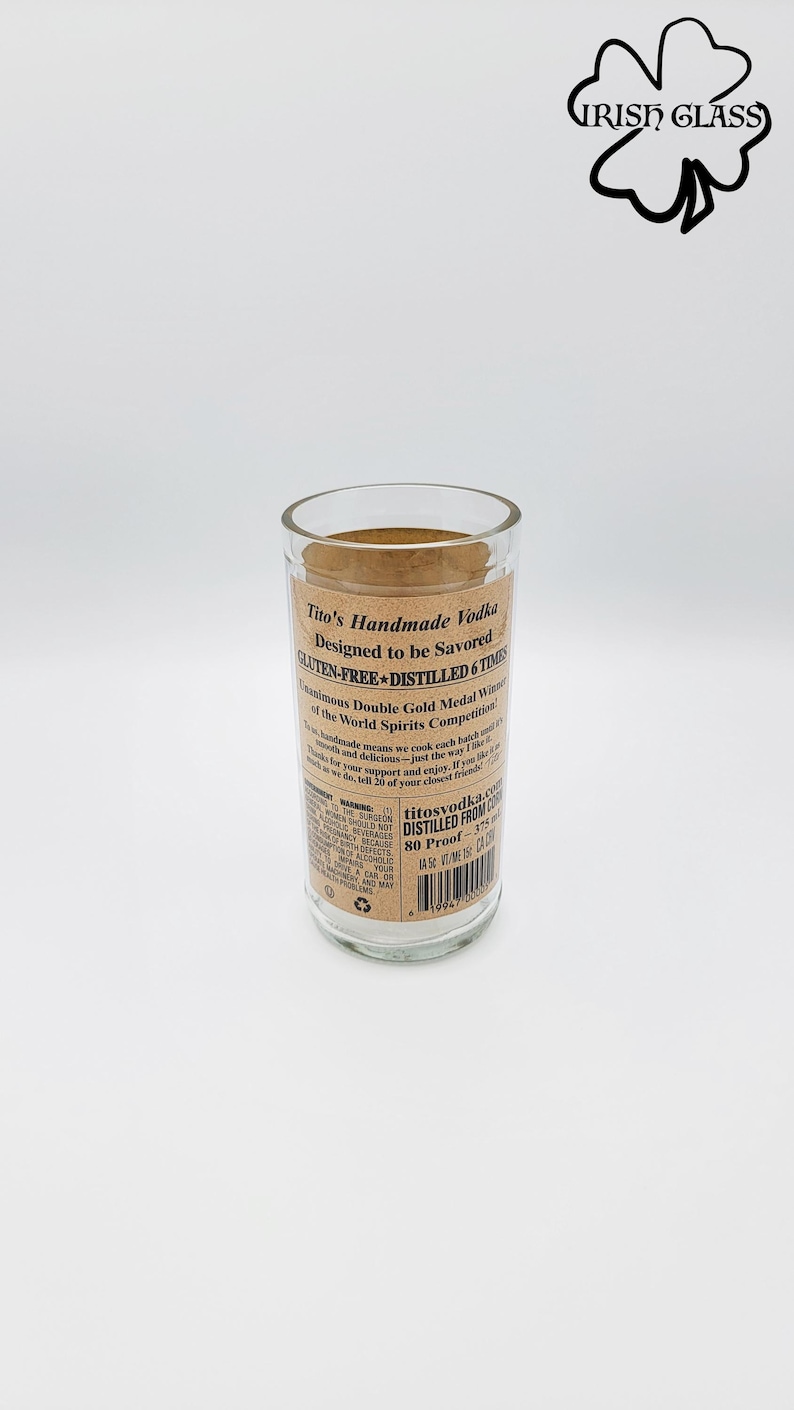 May include: A clear glass tumbler repurposed from a Tito's Handmade Vodka bottle. The label features black text on a tan background, stating Tito's Handmade Vodka, Gluten-Free, Distilled 6 Times, 80 Proof, and 375 ml. A shamrock logo reads Irish Glass.