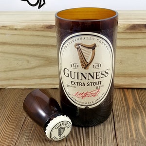 May include: A brown glass bottle with a white label that reads "Traditionally Brewed", "Est. 1759", "Guinness", "Extra Stout", "5.6% Alc./Vol.", "11.2 fl. oz.", "St. James's Gate Dublin". The bottle is sitting on a wooden surface. There is a smaller brown glass bottle with a white cap that reads "Guinness Extra Stout" on the wooden surface.
