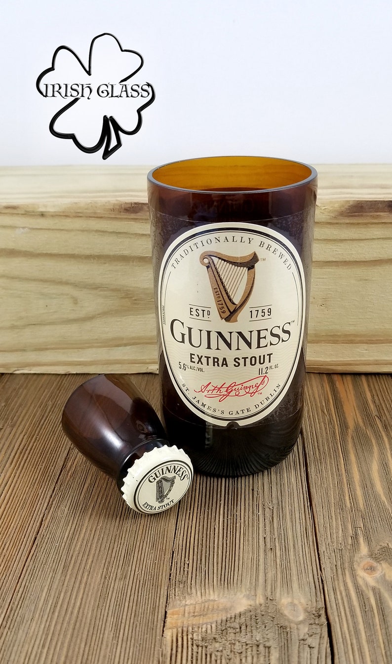 May include: A brown glass bottle with a white label that reads "Traditionally Brewed", "Est. 1759", "Guinness", "Extra Stout", "5.6% Alc./Vol.", "11.2 fl. oz.", "St. James's Gate Dublin". The bottle is sitting on a wooden surface. There is a smaller brown glass bottle with a white cap that reads "Guinness Extra Stout" on the wooden surface.