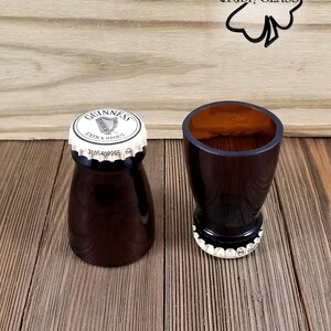 May include: Two brown glass shot glasses with a white Guinness cap on the top of each. The glasses are on a wooden surface with a shamrock and the words "Irish Glass" carved into the wood.