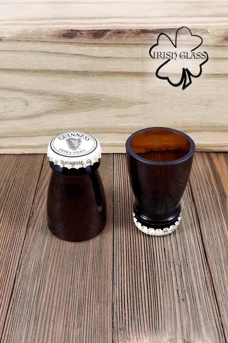 May include: Two brown glass shot glasses with a white Guinness cap on the top of each. The glasses are on a wooden surface with a shamrock and the words "Irish Glass" carved into the wood.