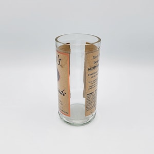 May include: A clear glass tumbler repurposed from a bottle, featuring a tan label with black text that reads Tito's Handmade Vodka, Gluten-Free, and includes a government warning. The top rim is smooth and the base is circular and transparent.